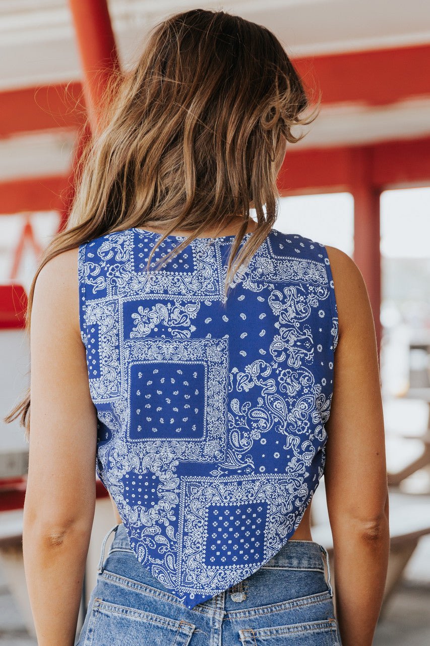 A woman with wavy hair wears the Out West Blue Bandana Print Sleeveless Cowl Neck Top and jeans outdoors, seen from behind.