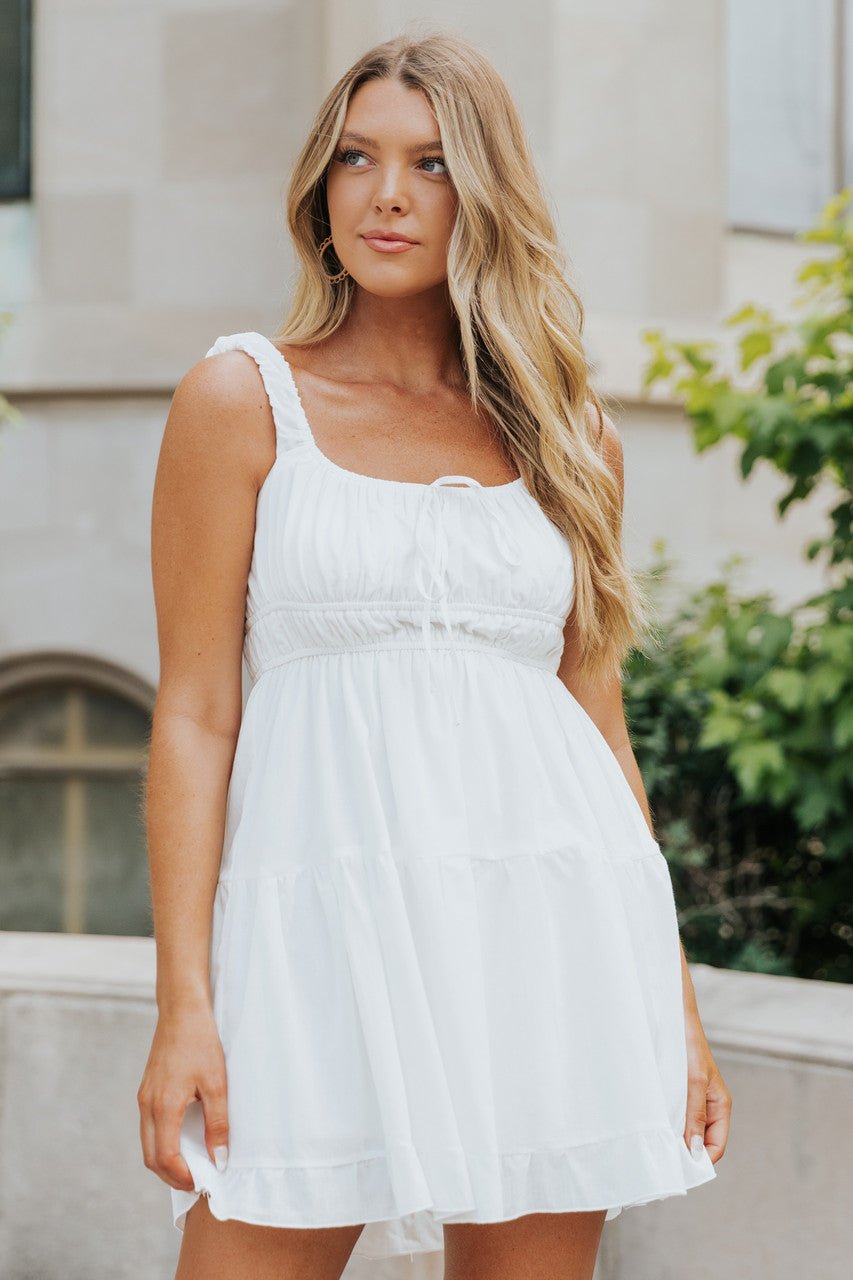 A woman with long blonde hair wears the Square Neck White Gathered Tiered Dress outdoors by a stone building and greenery.