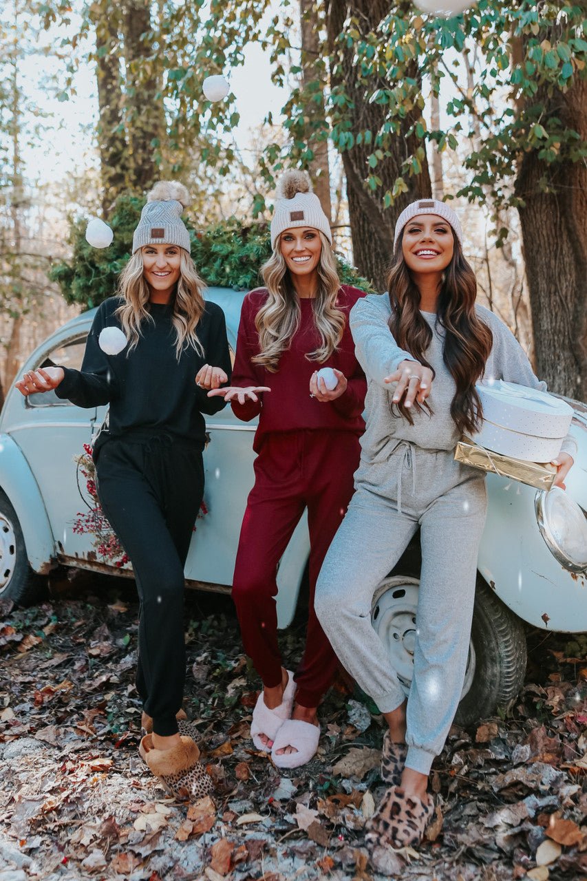 Three women in Step Up Lounge Set joggers and beanies stand by a vintage car with a Christmas tree, smiling and tossing snowballs.