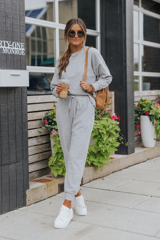 Woman in Step Up Lounge Set with white sneakers, sunglasses, and tan backpack holds coffee by plants outside a building.