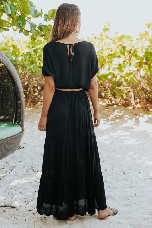 Woman on sandy ground in the Sunrise Short Sleeve Top & Maxi Skirt Black Set, facing away with greenery behind her.