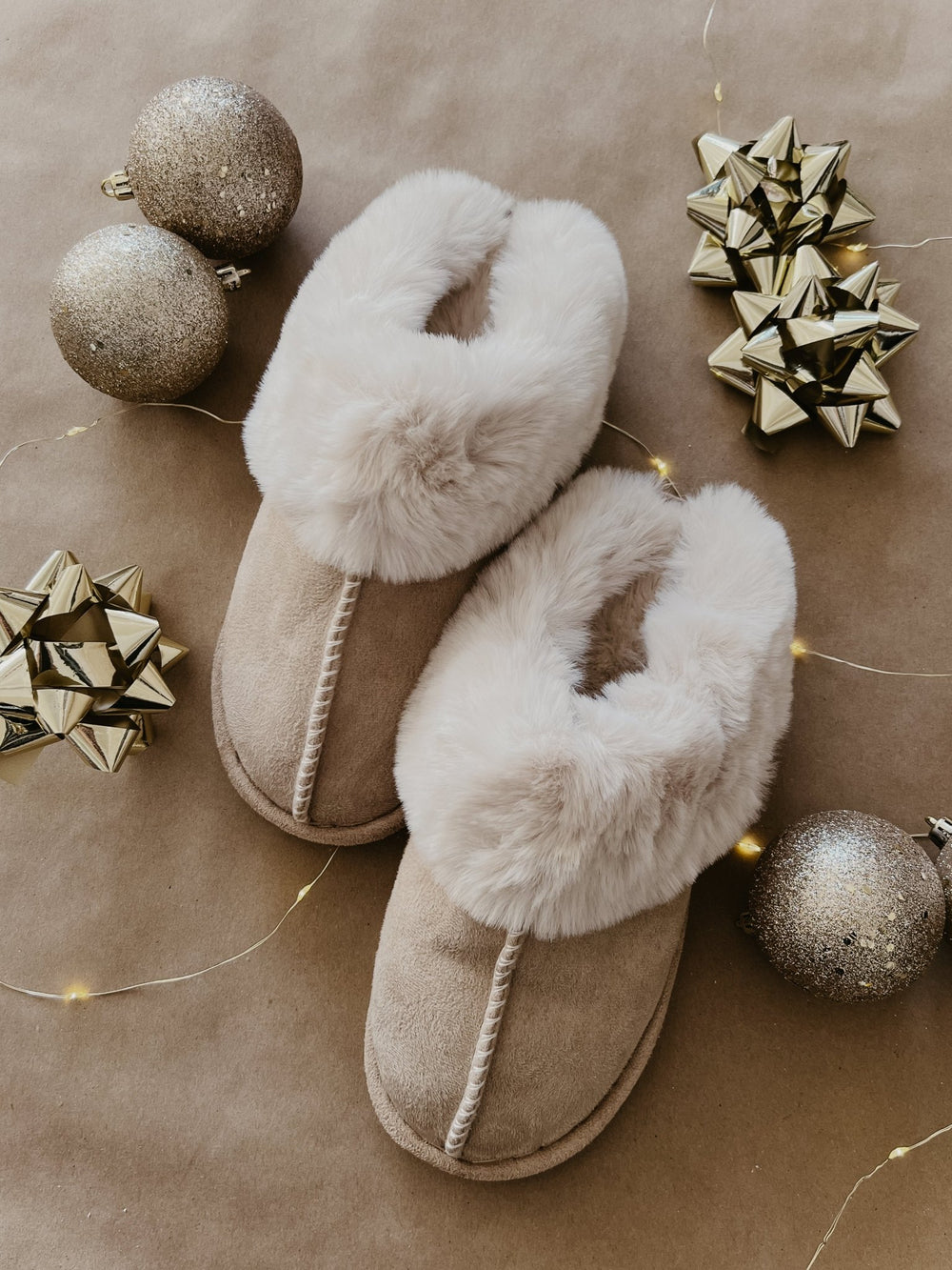 Sweet Dreams Sandy Faux Sheepskin Slippers in beige, surrounded by gold ornaments and gift bows on a brown background.