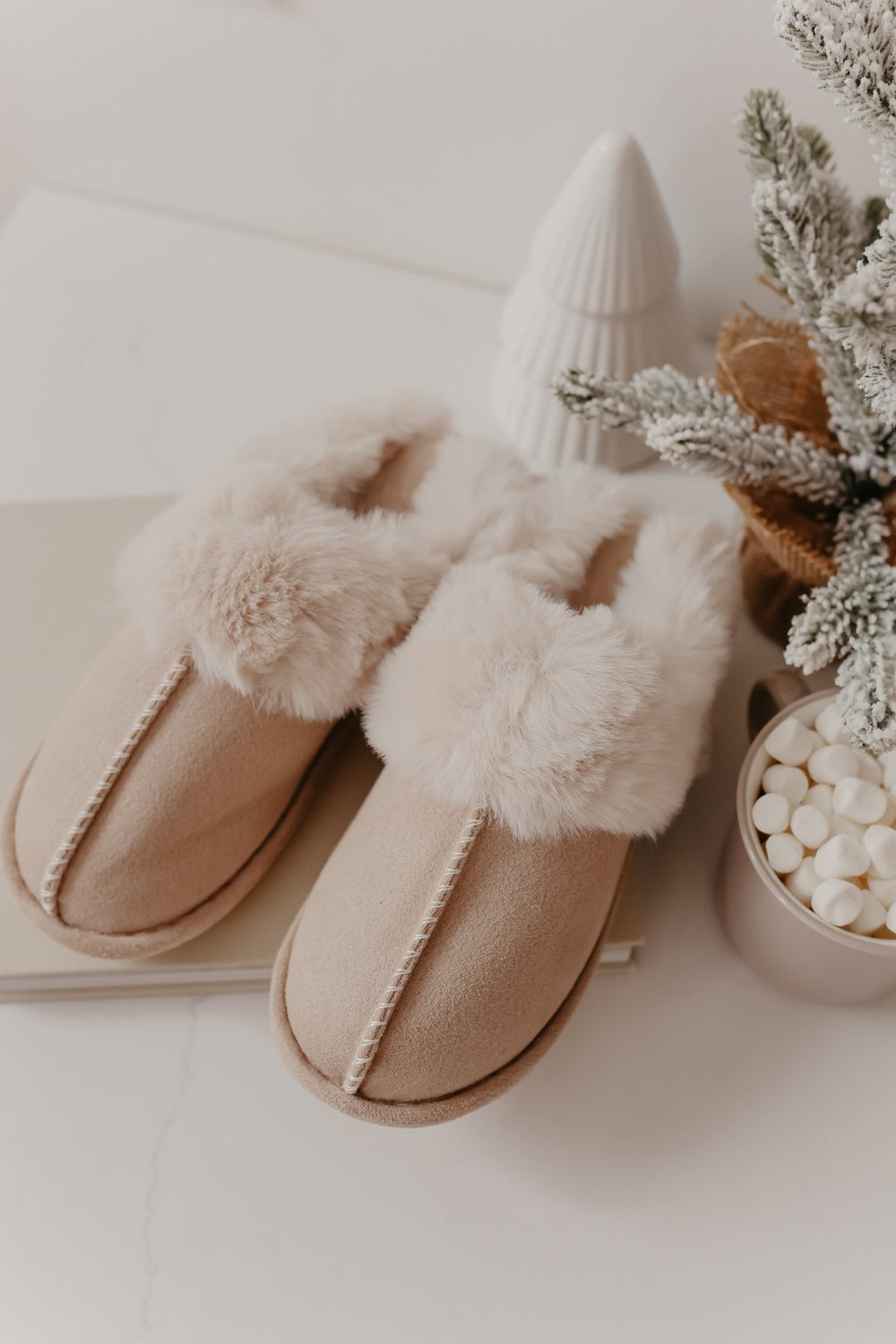 The Sweet Dreams Sandy Faux Sheepskin Slippers in beige are displayed beside a mug of marshmallows and a snowy holiday decor.