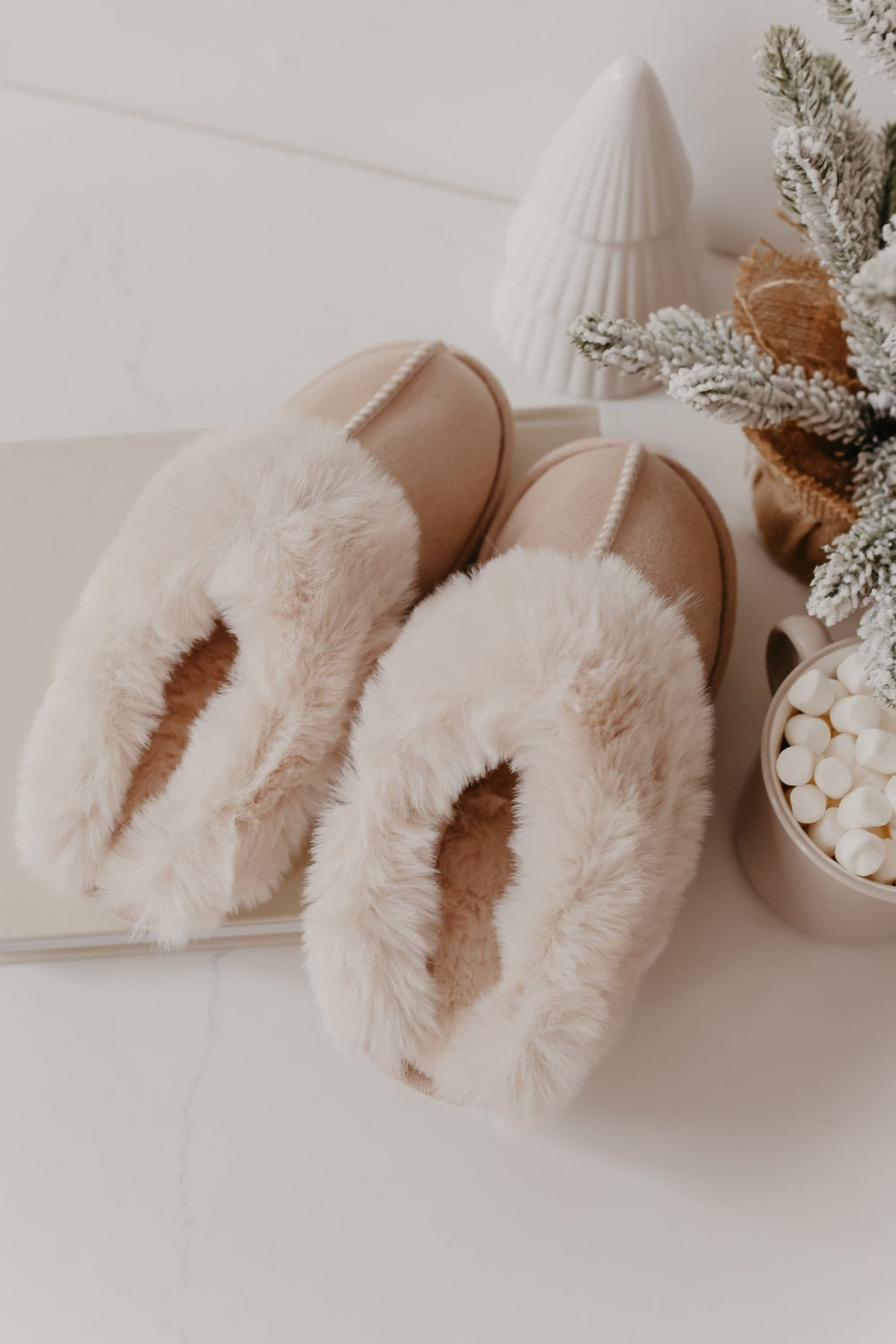 Sweet Dreams Sandy Faux Sheepskin Slippers beside a marshmallow cup, winter plant arrangement, and white ceramic tree.