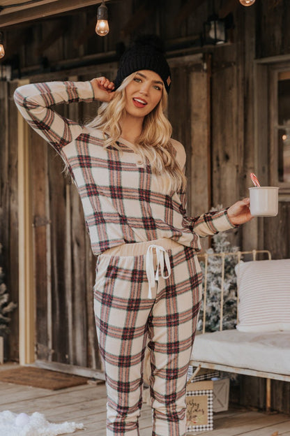 Woman in Taupe Tartan Plaid Jogger Pajama Pants - FINAL SALE smiles, holding a mug on a festive winter porch.