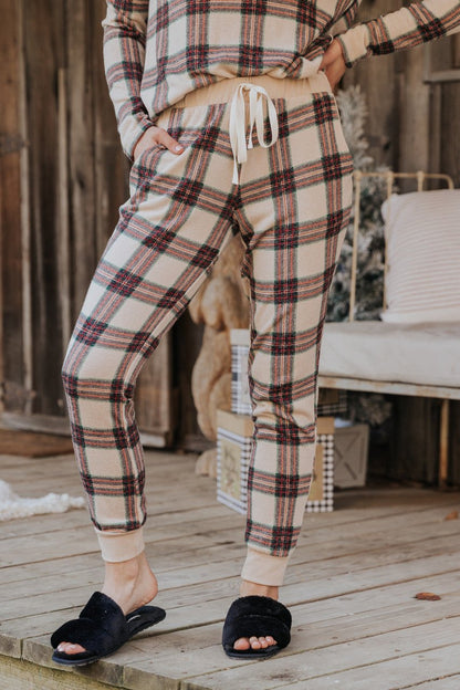 Wearing Taupe Tartan Plaid Jogger Pajama Pants - FINAL SALE and black slippers, a person stands on a wooden porch with hands in pockets.