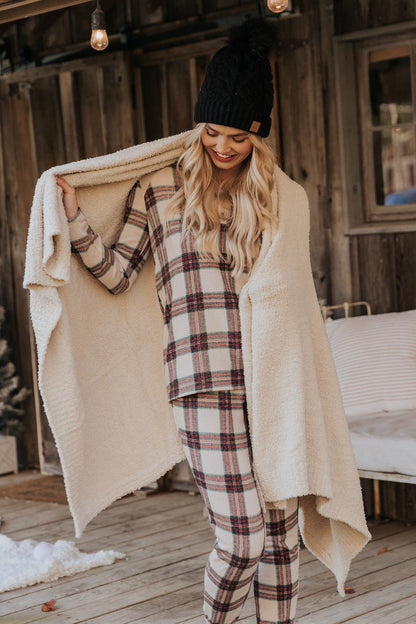 A woman in Taupe Tartan Plaid Jogger Pajama Pants and a black knit hat smiles on a porch, wrapped in a cozy beige blanket.