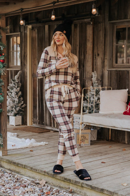 A woman in Taupe Tartan Plaid Jogger Pajama Pants - FINAL SALE holds a mug on a holiday-decorated porch.