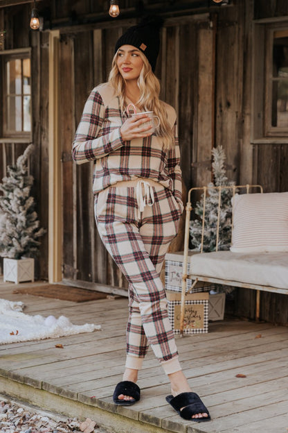 Woman in taupe tartan plaid jogger pajama pants stands on a wooden porch, holding a mug, with snowy decor in the background.