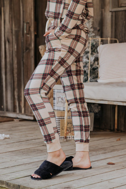 Person wearing Taupe Tartan Plaid Jogger Pajama Pants - FINAL SALE stands on a porch, one foot raised, hands in pockets.