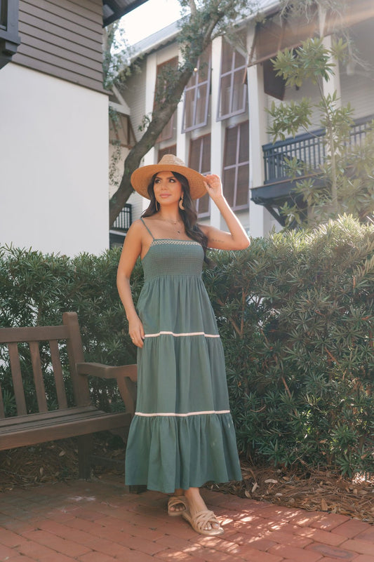 Woman in a Teal Smocked Cami Maxi Dress and straw hat stands outdoors by greenery and a bench, houses in the background.