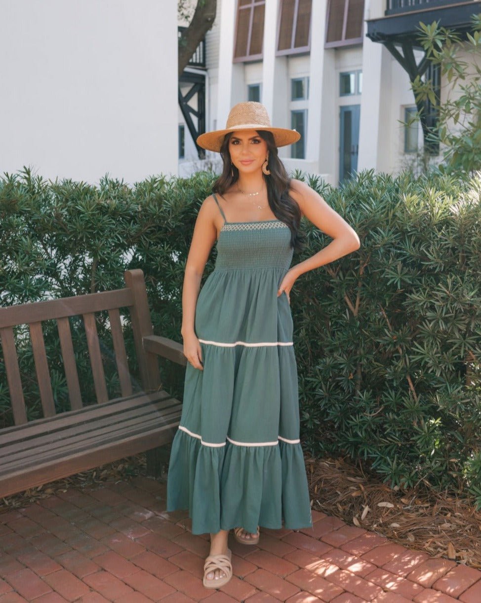 Woman in a Teal Smocked Cami Maxi Dress and straw hat poses outdoors by a wooden bench and greenery on a sunny brick path.