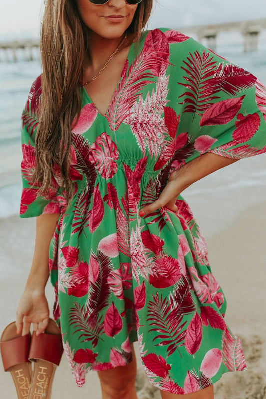 Woman in a V-Neck Green/Pink Palm Empire Dress stands on a beach holding sandals, with waves and a pier in the background.
